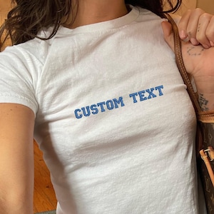 May include: White cropped t-shirt with blue text that reads "CUSTOM TEXT". The shirt is being worn by a person with a brown leather handbag with a gold chain.