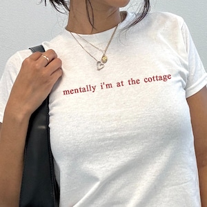 May include: A white short-sleeved t-shirt featuring the red text "mentally i'm at the cottage" printed on the chest. The shirt is paired with a black shoulder bag and a black skirt with a red floral pattern, accessorised with a gold heart necklace.