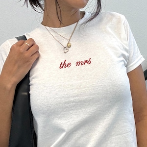 May include: White t-shirt with the text "the mrs" in red script. The shirt is short-sleeved and appears to be made of a soft material. The person is wearing black sunglasses and a black handbag.