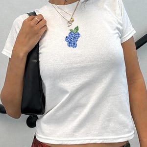 May include: A white short-sleeved t-shirt featuring a small graphic of blue chequered grapes with green leaves centred on the chest. The shirt is paired with a black shoulder bag and a black skirt with a red cherry print pattern.