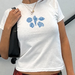 May include: A white short-sleeved t-shirt featuring a central graphic print of five blue seashells arranged in a cluster. The shirt has a crew neckline and is worn with a black shoulder bag, a layered gold necklace, and black sunglasses.