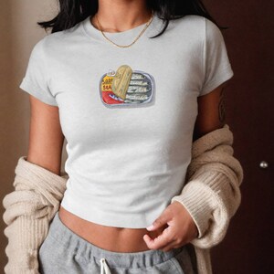 May include: A white t-shirt featuring a graphic of a sardine can. The can is open and the sardines are visible inside. The text on the can reads "SARDINES SEA".