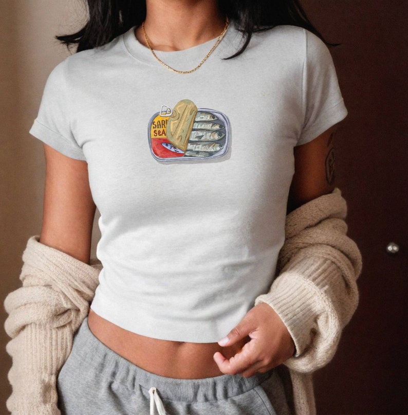 May include: A white t-shirt featuring a graphic of a sardine can. The can is open and the sardines are visible inside. The text on the can reads "SARDINES SEA".