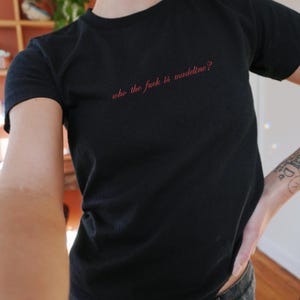 May include: Black t-shirt with the red text "who the fuck is madeline?" The shirt is short-sleeved and appears to be made of a soft material. The text is centered on the chest.