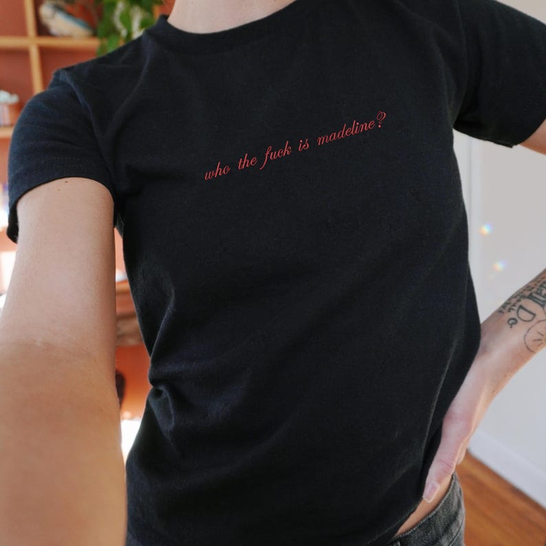 May include: Black t-shirt with the red text "who the fuck is madeline?" The shirt is short-sleeved and appears to be made of a soft material. The text is centered on the chest.