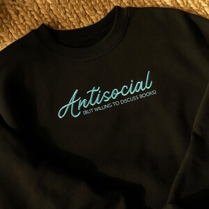May include: Black sweatshirt with the word "Antisocial" embroidered in a light blue script font. Below the word "Antisocial" is the phrase "(BUT WILLING TO DISCUSS BOOKS)" in a smaller font.