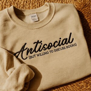 May include: Beige sweatshirt with the word "Antisocial" embroidered in black script, with the phrase "(BUT WILLING TO DISCUSS BOOKS)" below. The Gildan tag is visible. The sweatshirt is folded on a woven surface.