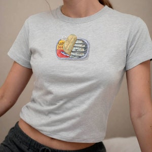 May include: A light grey t-shirt featuring a graphic of a tin of sardines. The tin is open and the sardines are visible inside. The tin has the text "SARDINES SEA" on it.