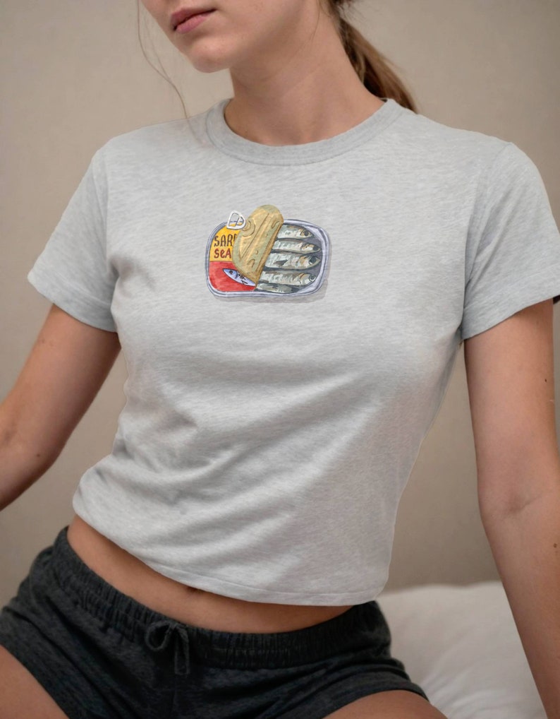May include: A light grey t-shirt featuring a graphic of a tin of sardines. The tin is open and the sardines are visible inside. The tin has the text "SARDINES SEA" on it.