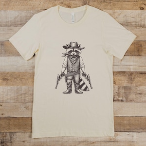 May include: A cream-coloured t-shirt featuring a graphic of a raccoon dressed as a cowboy. The raccoon is wearing a hat, vest, and holding two pistols. The raccoon is standing with its legs spread apart and has a bushy tail.