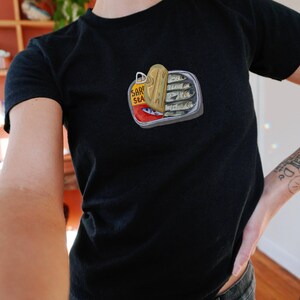 May include: A black t-shirt featuring a graphic of a sardine can with the text "Sard Sea" on it.