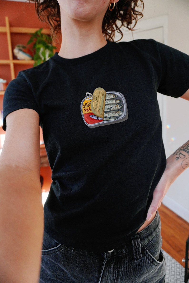 May include: A black t-shirt featuring a graphic of a sardine can with the text "Sard Sea" on it.