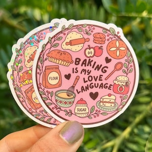 May include: A round sticker with a pink background featuring illustrations of baking items like a flour bag, whisk, pie, and cake. The text reads Baking is my love language, surrounded by a decorative border of leaves and small hearts.