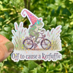 May include: A whimsical sticker featuring a green frog riding a bicycle, wearing a pink and white wizard hat. The sticker has the text "Off to cause a Kerfuffle" and is surrounded by decorative elements.