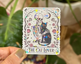 May include: A sticker featuring a skeleton embracing a black cat, with a crescent moon and stars. The sticker has a floral border with the text "THE CAT LOVER". The design has a holographic effect.