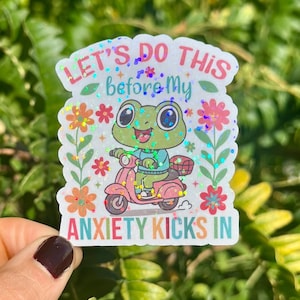 May include: A vibrant sticker showing a cartoon frog on a pink scooter, with the text "LET'S DO THIS before My ANXIETY KICKS IN". The design includes floral and glitter elements. The sticker is set against a green backdrop.