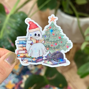 May include: A whimsical sticker featuring a ghost wearing a Santa hat, reading a book, with a Christmas tree and a sleeping black cat. The ghost is surrounded by stacks of books, creating a cozy, festive scene.