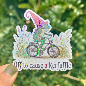 May include: A whimsical sticker featuring a frog riding a bicycle, wearing a wizard hat. The frog is green with blue eyes. The sticker has the text "Off to cause a Kerfuffle". The background has glitter and grass.