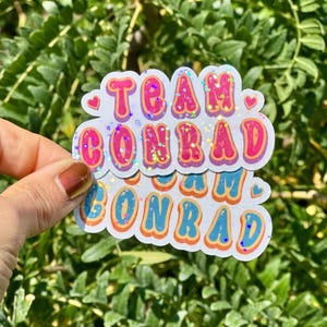 May include: Two stickers with the text "TEAM CONRAD" in a retro font. The top sticker has pink and orange letters with glitter accents, and the bottom sticker has blue and orange letters. Both stickers have a white border and heart details.