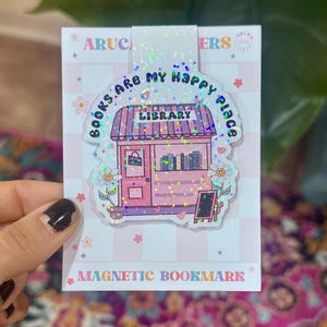 May include: A magnetic bookmark featuring a holographic finish and a pink library illustration. The text reads "BOOKS ARE MY HAPPY PLACE" and "LIBRARY". The design includes flowers with smiley faces, a door, and a small chalkboard sign.