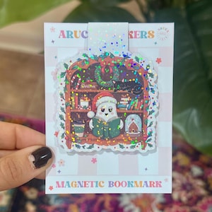 May include: A magnetic bookmark on a white and light pink checkered backing card with the text ARUCA STICKERS and MAGNETIC BOOKMARK. The bookmark features a holographic ghost wearing a Santa hat, reading a book in a wooden bookcase with a cat.