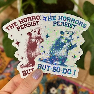 May include: Two iridescent stickers with a possum graphic and the text "The Horrors Persist But So Do I".