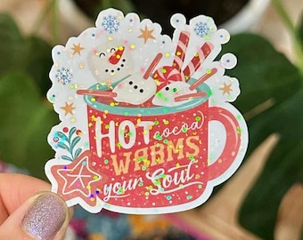 May include: A colorful sticker featuring a mug of hot cocoa with a snowman and candy canes. The text "Hot cocoa warms your soul" is displayed on the mug. The sticker has glitter accents and a star-shaped element.