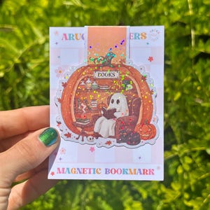 May include: A magnetic bookmark featuring a holographic pumpkin-shaped illustration. Inside the pumpkin, a white ghost sits in a brown armchair reading a book. The scene includes shelves with jars, books, a small skull, and a jack-o'-lantern. The text reads BOOKS.