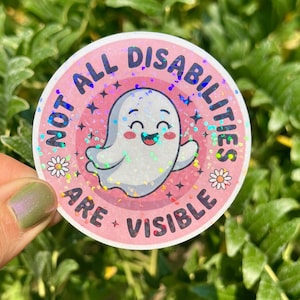 May include: A round sticker with the text "NOT ALL DISABILITIES ARE VISIBLE" in pink and white. It features a smiling ghost illustration with a holographic effect, surrounded by stars and daisies.