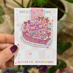 May include: A pink magnetic bookmark with a heart-shaped cake design. The cake has the words "Hot Girls Read Books" and other illustrations. The bookmark is held by a hand with a dark purple nail polish.