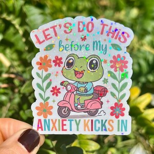 May include: A holographic sticker with a cartoon frog on a pink scooter. The text reads "LET'S DO THIS before My ANXIETY KICKS IN", with floral and glitter details. The frog is green and wearing a blue sweater.