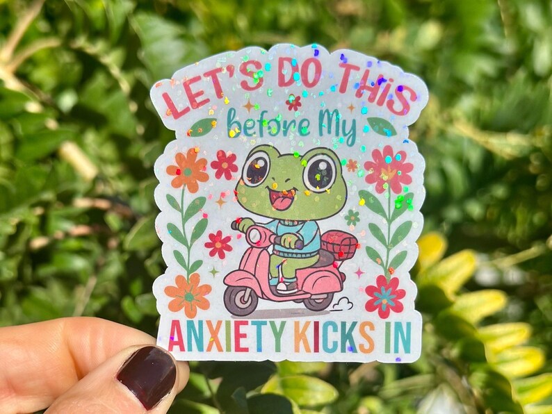 May include: A holographic sticker with a cartoon frog on a pink scooter. The text reads "LET'S DO THIS before My ANXIETY KICKS IN", with floral and glitter details. The frog is green and wearing a blue sweater.
