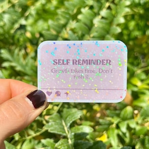 May include: A rectangular sticker with rounded corners, featuring the text "SELF REMINDER" and the message "Growth takes time. Don't rush it." The sticker has a glittery, iridescent effect with small icons at the bottom.