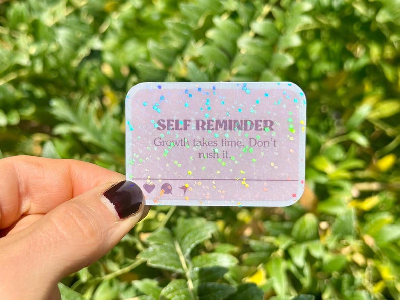 May include: A rectangular sticker with rounded corners, featuring the text "SELF REMINDER" and the message "Growth takes time. Don't rush it." The sticker has a glittery, iridescent effect with small icons at the bottom.