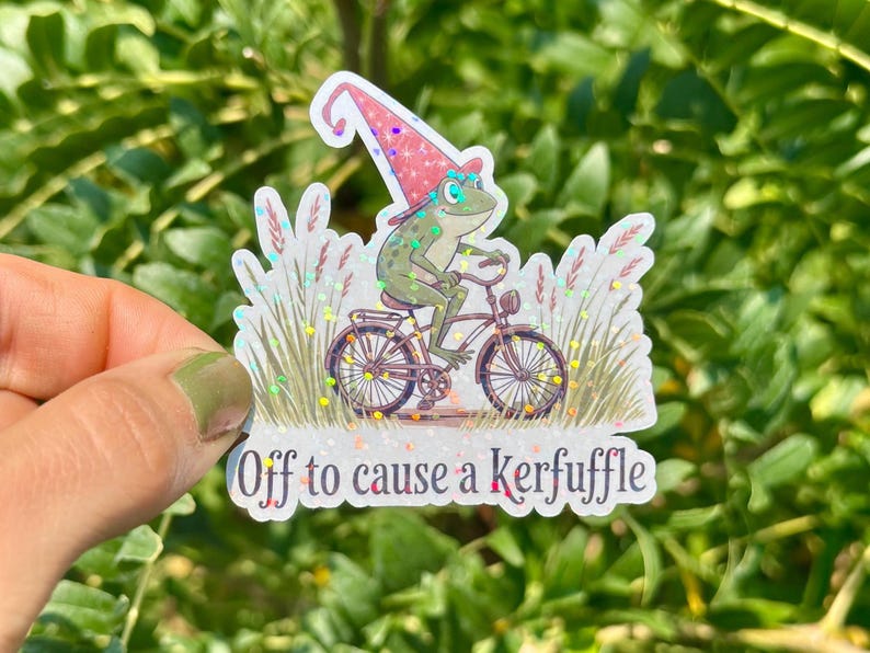 May include: A whimsical sticker featuring a green frog riding a bicycle, wearing a pink and white wizard hat. The sticker has the text "Off to cause a Kerfuffle" and is surrounded by decorative elements.