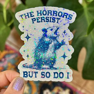 May include: A white sticker with a blue opossum illustration and the text "The Horrors Persist, But So Do I". The sticker has a glittery, iridescent finish.