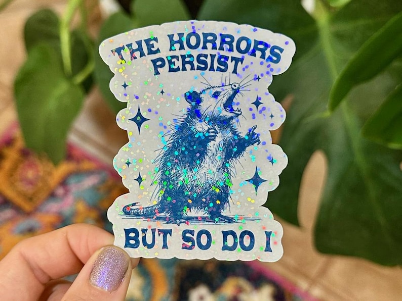 May include: A white sticker with a blue opossum illustration and the text "The Horrors Persist, But So Do I". The sticker has a glittery, iridescent finish.
