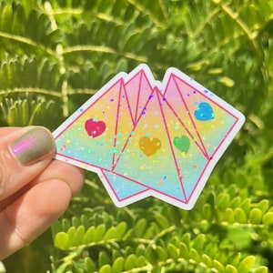 May include: A holographic sticker with a rainbow gradient and three hearts in different colors. The sticker is shaped like three overlapping triangles.