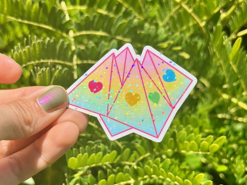 May include: A holographic sticker with a rainbow gradient and three hearts in different colors. The sticker is shaped like three overlapping triangles.