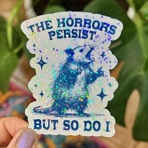 May include: A white sticker with a blue opossum illustration and the text "The horrors persist but so do I". The sticker has a glittery finish.