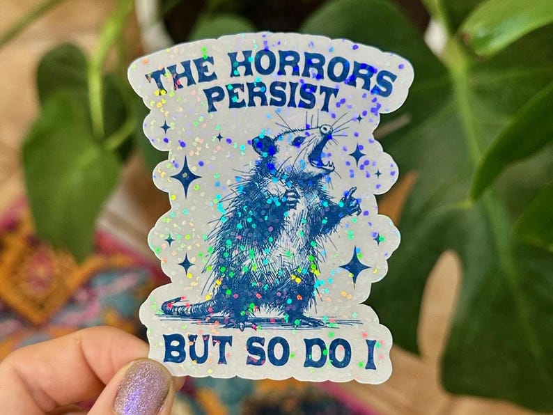 May include: A white sticker with a blue opossum illustration and the text "The horrors persist but so do I". The sticker has a glittery finish.