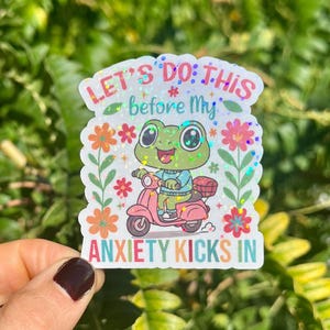 May include: A sticker with a cartoon frog on a pink scooter. The text reads "LET'S DO THIS before My ANXIETY KICKS IN", with floral and glitter details. The frog wears a blue top and the scooter has a basket. The sticker is white.