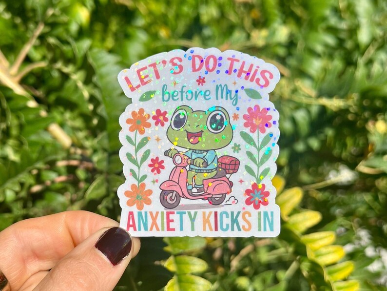 May include: A sticker with a cartoon frog on a pink scooter. The text reads "LET'S DO THIS before My ANXIETY KICKS IN", with floral and glitter details. The frog wears a blue top and the scooter has a basket. The sticker is white.