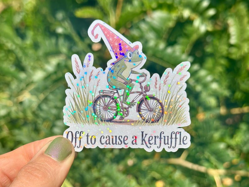 May include: A whimsical sticker featuring a frog riding a bicycle, wearing a wizard hat. The frog is green with blue eyes. The sticker has the text "Off to cause a Kerfuffle". The background has glitter and grass.