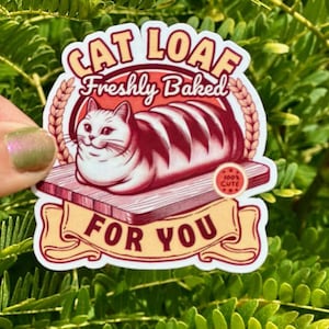 May include: A sticker with a cartoon cat shaped like a loaf of bread. The cat is white with pink and brown stripes. The sticker says "Cat Loaf Freshly Baked For You" and has a small circle that says "100% Cute".