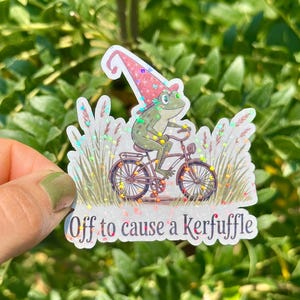 May include: A whimsical sticker featuring a frog in a gnome hat riding a bicycle. The frog is green with a red and pink hat. The sticker has the text "Off to cause a kerfuffle".