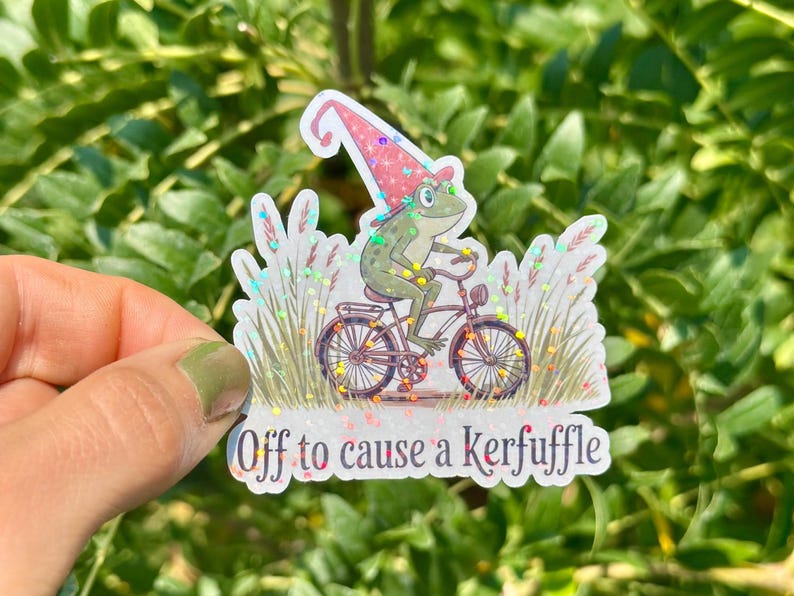 May include: A whimsical sticker featuring a frog in a gnome hat riding a bicycle. The frog is green with a red and pink hat. The sticker has the text "Off to cause a kerfuffle".