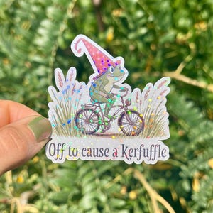 May include: A whimsical sticker featuring a green frog riding a bicycle, wearing a pink and white wizard hat. The sticker has the text "Off to cause a kerfuffle" and is surrounded by decorative grass.