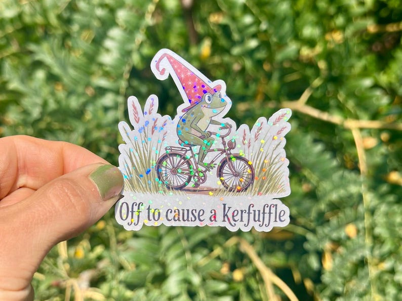 May include: A whimsical sticker featuring a green frog riding a bicycle, wearing a pink and white wizard hat. The sticker has the text "Off to cause a kerfuffle" and is surrounded by decorative grass.