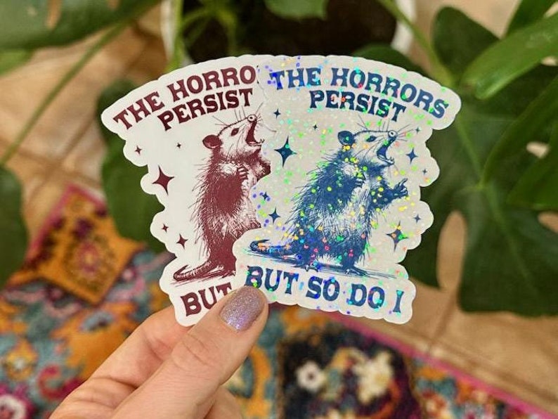 May include: Two iridescent stickers with a possum graphic and the text "The Horrors Persist But So Do I".